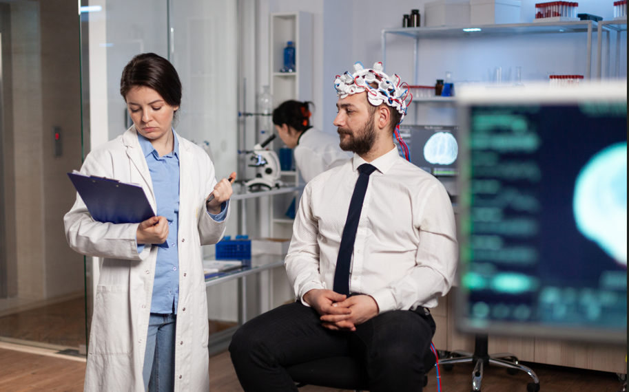 Female doctor reviewing charts of a patient with brain device on while sitting on a stool.