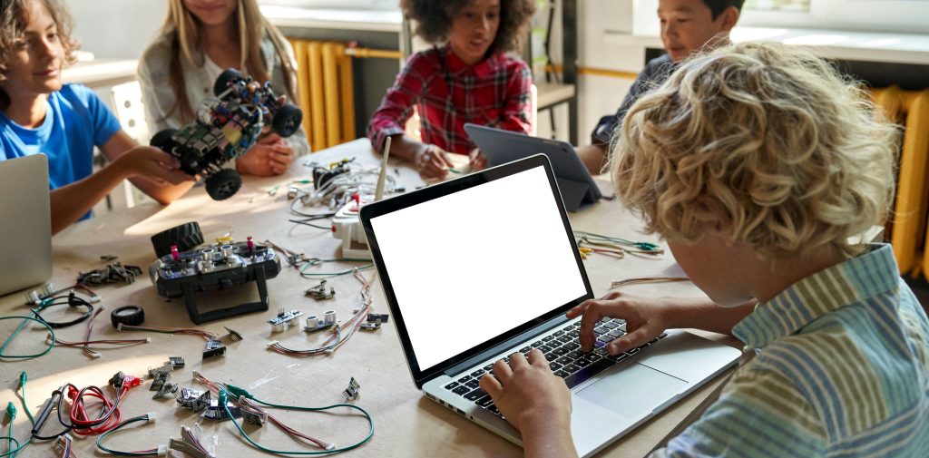 A student works at a laptop alongside classmates building and coding robotics projects at a STEM table.