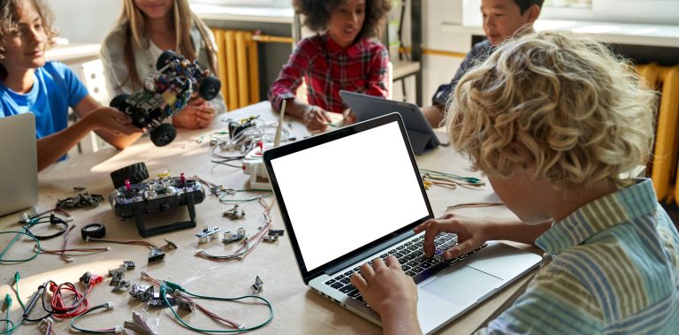 A student works at a laptop alongside classmates building and coding robotics projects at a STEM table.